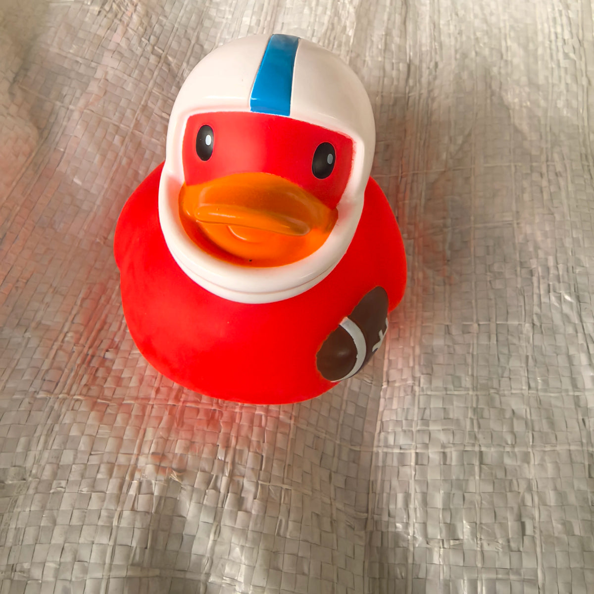A bright red rubber duck bath toy with an orange beak and blue stripe sitting on a silver textured surface.