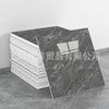 A rectangular slab of dark gray faux marble material is shown next to a stack of white tiles, demonstrating the peel-and-stic