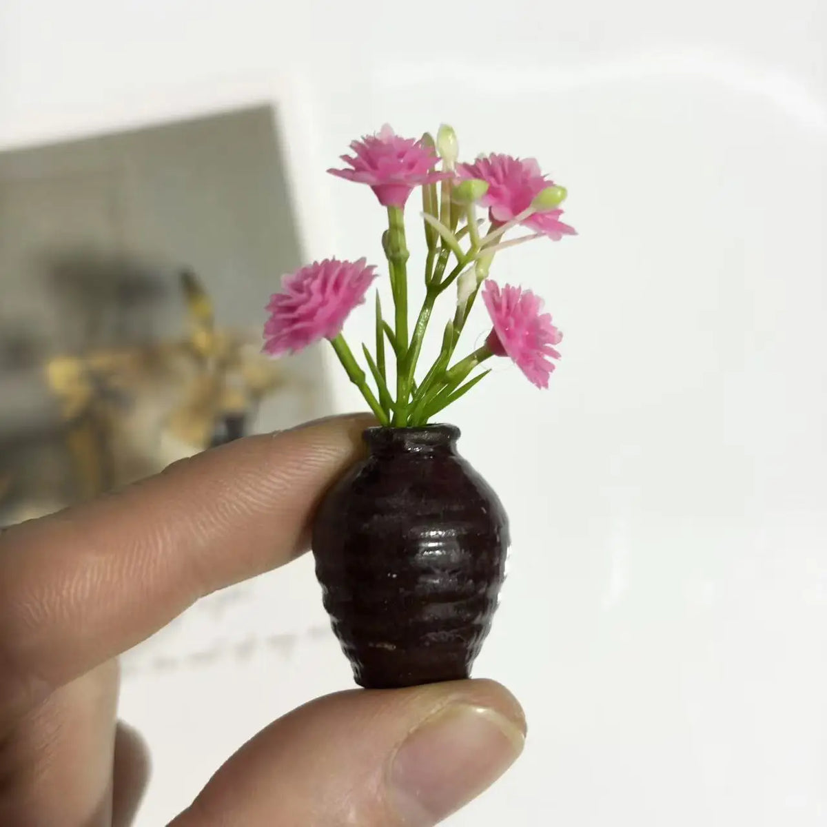 A hand holds a dark brown ceramic vase with pink artificial flowers, used as a fridge magnet.