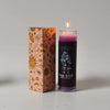 A glass candle in a tall cylinder with purple wax and a lit wick, next to its decorative cardboard box.