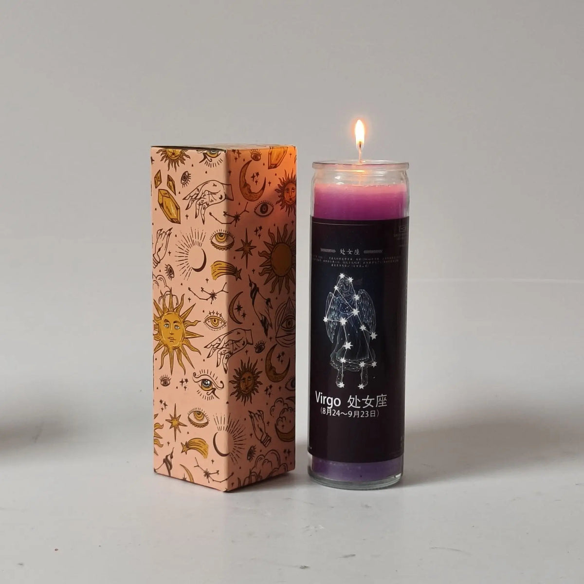 A glass candle in a tall cylinder with purple wax and a lit wick, next to its decorative cardboard box.