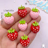 A hand holds various colorful strawberry resin flatback charms in shades of red, pink, and green for DIY crafts.