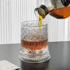 A rotating crystal whiskey glass with a relief design is being filled with amber liquid from a bottle.