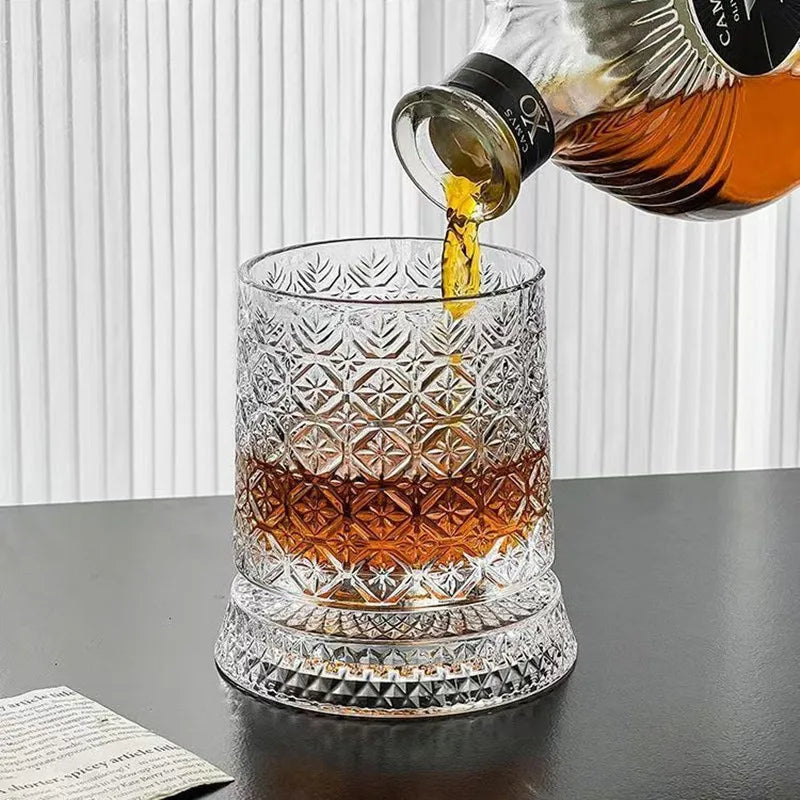 A rotating crystal whiskey glass with a relief design is being filled with amber liquid from a bottle.