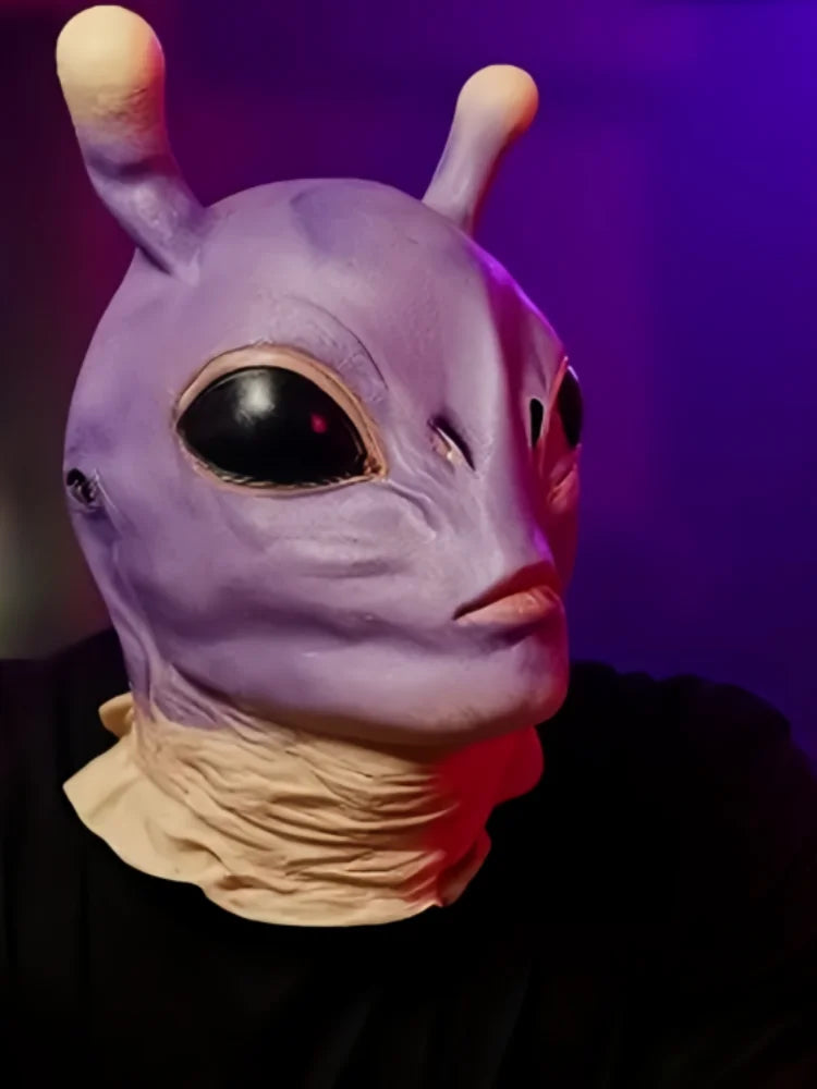A close-up of a purple alien full-face latex mask with large black eyes and pale skin, worn by a person.