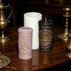 Three candles, one white and two dark, sit on a wooden surface next to a brass oil lamp.