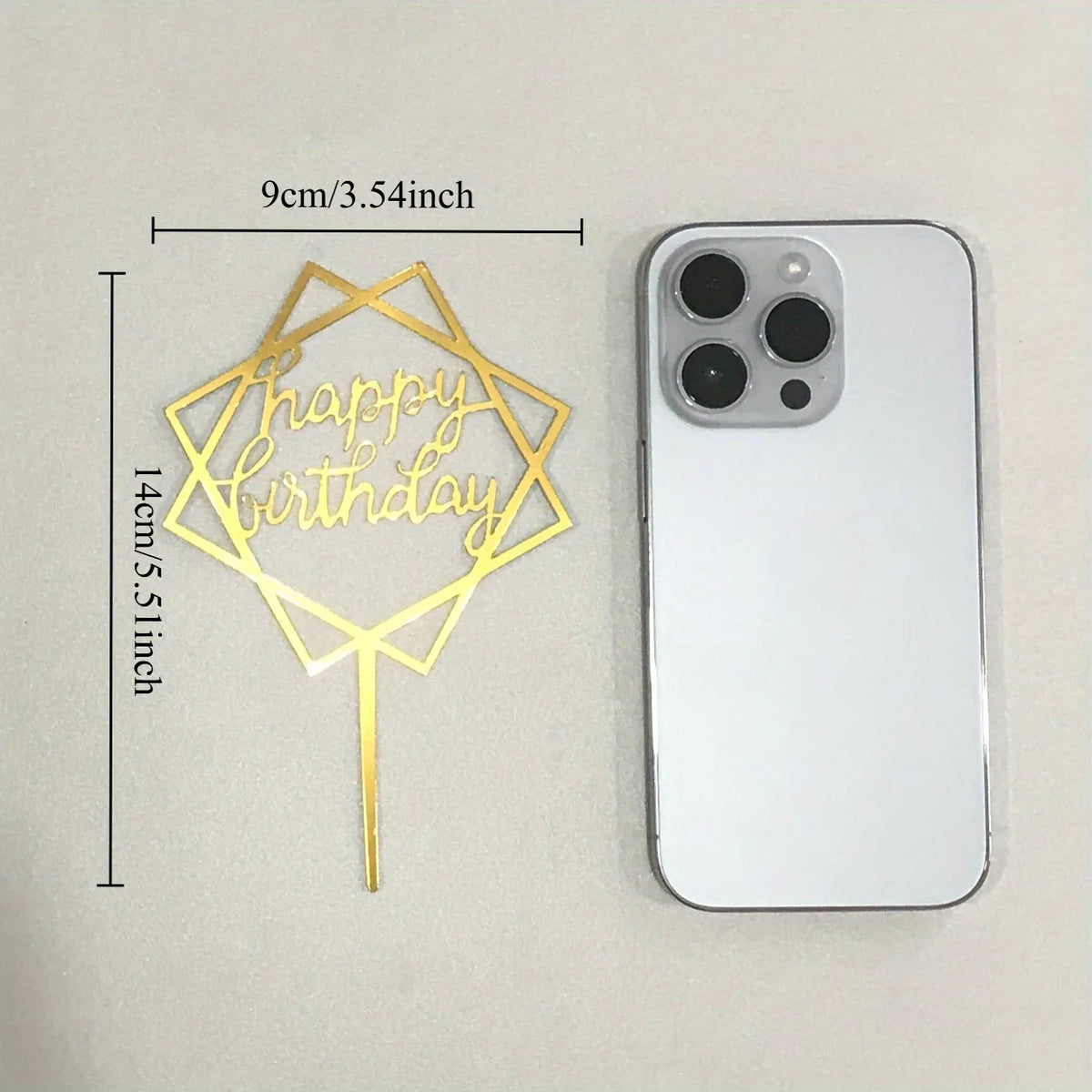 A gold acrylic happy birthday cake topper with a stick, shown next to an iPhone.