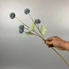 Una mano sostiene un tallo con flores artificiales de crisantemo y diente de león azul gris contra un fondo gris claro.