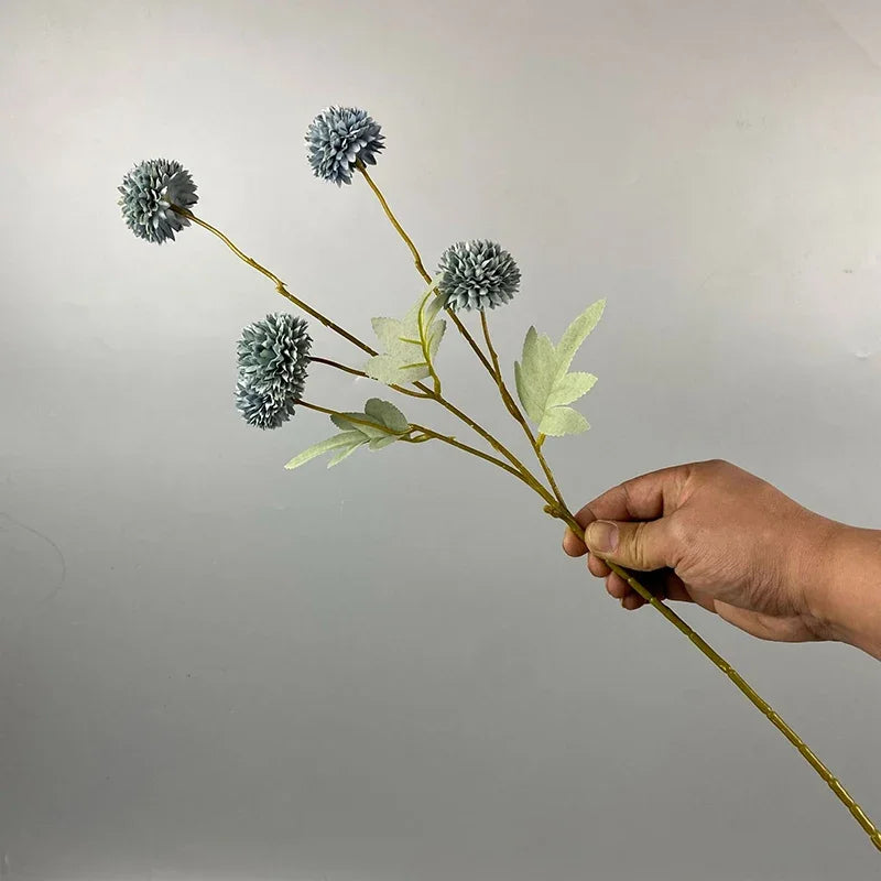 Una mano sostiene un tallo con flores artificiales de crisantemo y diente de león azul gris contra un fondo gris claro.