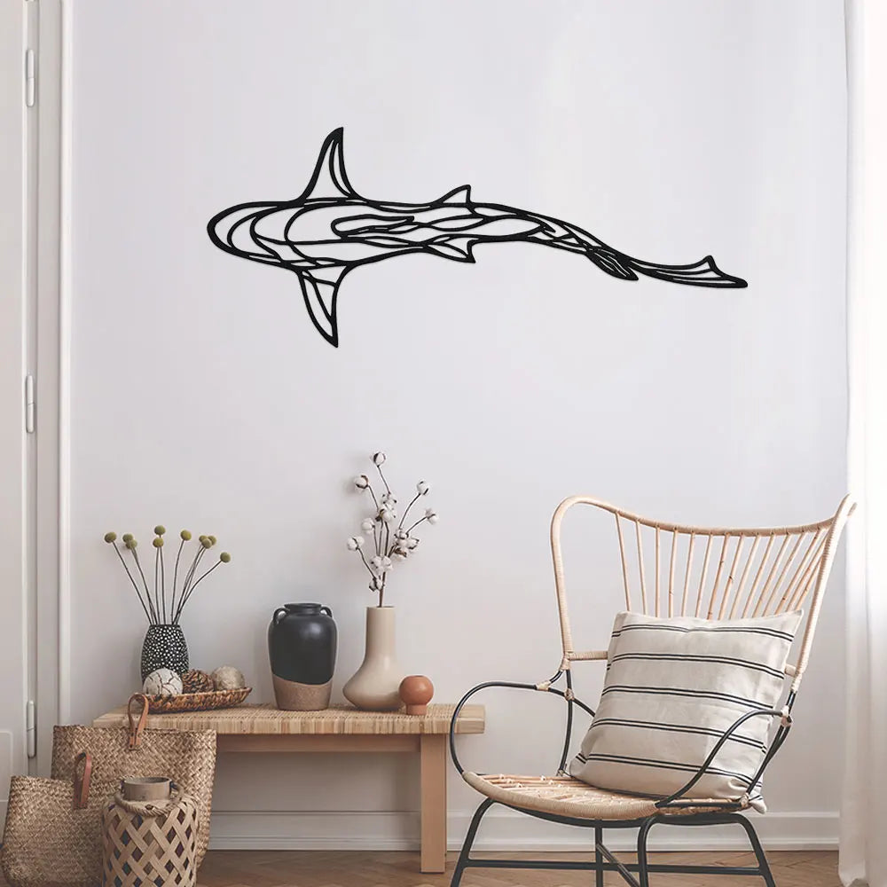 A black line sculpture of a shark swimming across a white wall above a wooden table and wicker chair.