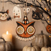 A wooden owl hanging ornament with patterned wings suspended from a dark branch against a blurred background of pumpkins and 