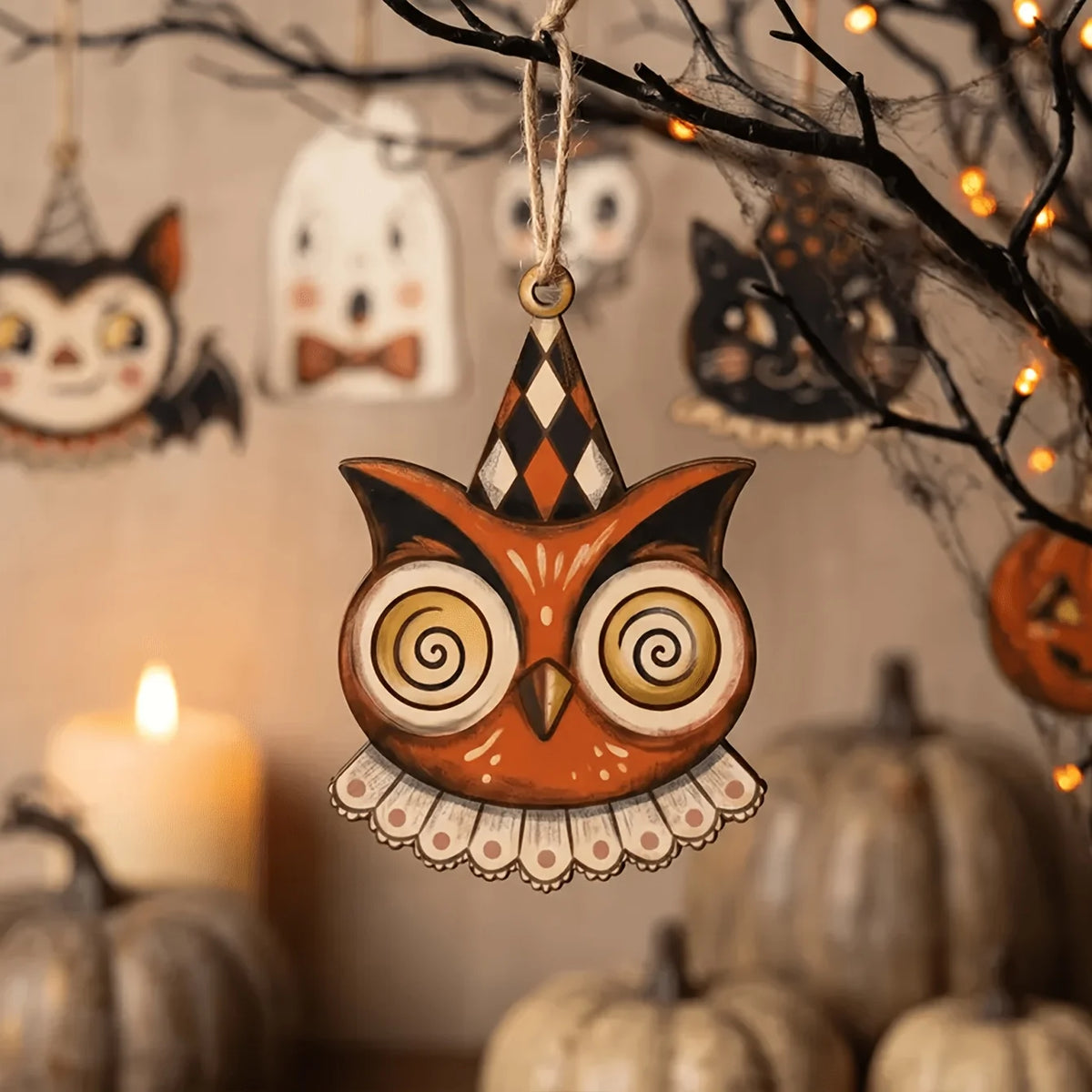 A wooden owl hanging ornament with patterned wings suspended from a dark branch against a blurred background of pumpkins and 