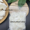 A clear plastic bag contains white natural coconut wax pieces next to a wooden board and green leaves.