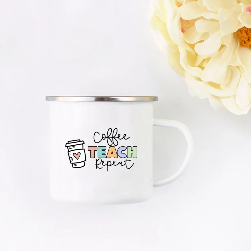 A white enamel mug with black text that reads Coffee Teach Repeat sits on a white surface next to a cluster of pale yellow fl
