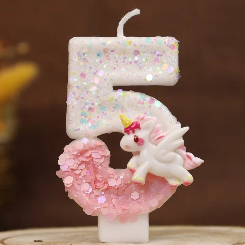 Una vela blanca con forma de 5, brillo rosa y una pequeña figura de unicornio blanco.