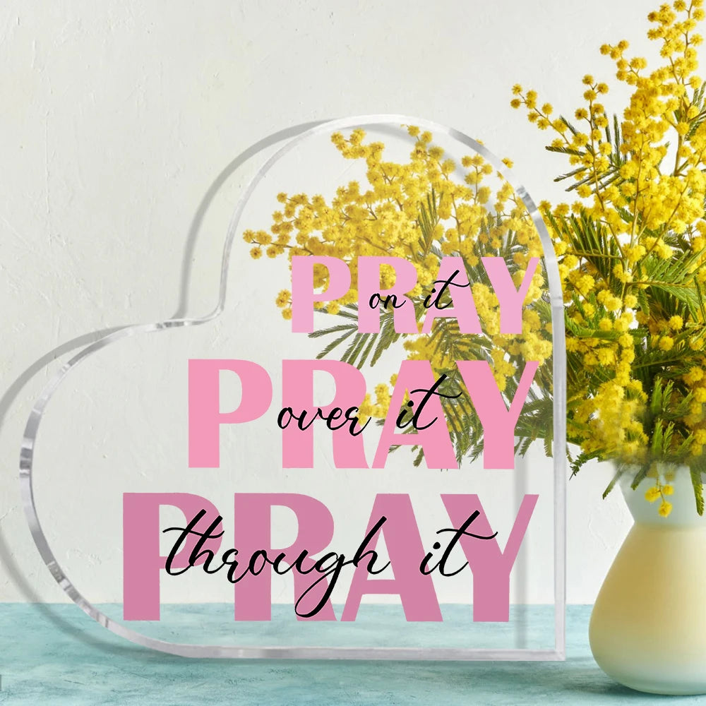 A clear acrylic heart-shaped sign with the text Pray it over it pray through it overlaid on a background of yellow flowers.