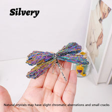 A hand holds a silvery crystal dragonfly figurine with iridescent colors against a white background.