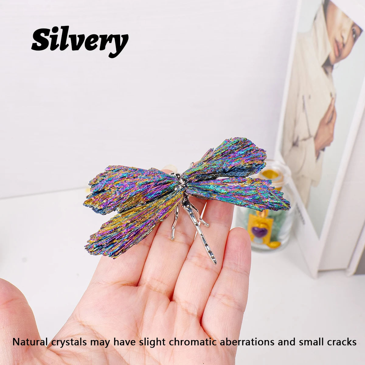 A hand holds a silvery crystal dragonfly figurine with iridescent colors against a white background.