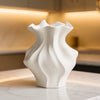 A white ceramic vase with a textured wave pattern sits on a marble surface in a softly lit interior.