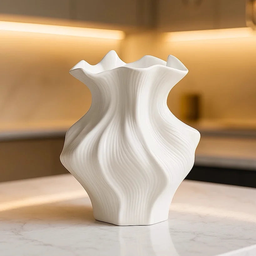 A white ceramic vase with a textured wave pattern sits on a marble surface in a softly lit interior.