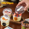A hand holds a clear reusable pudding cup filled with layered dessert, next to other cups and a wooden board.