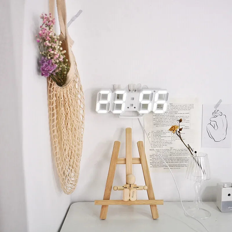 ساعة حائط رقمية ثلاثية الأبعاد LED بيضاء معروضة على رف بجانب حامل خشبي وبعض الزهور المجففة