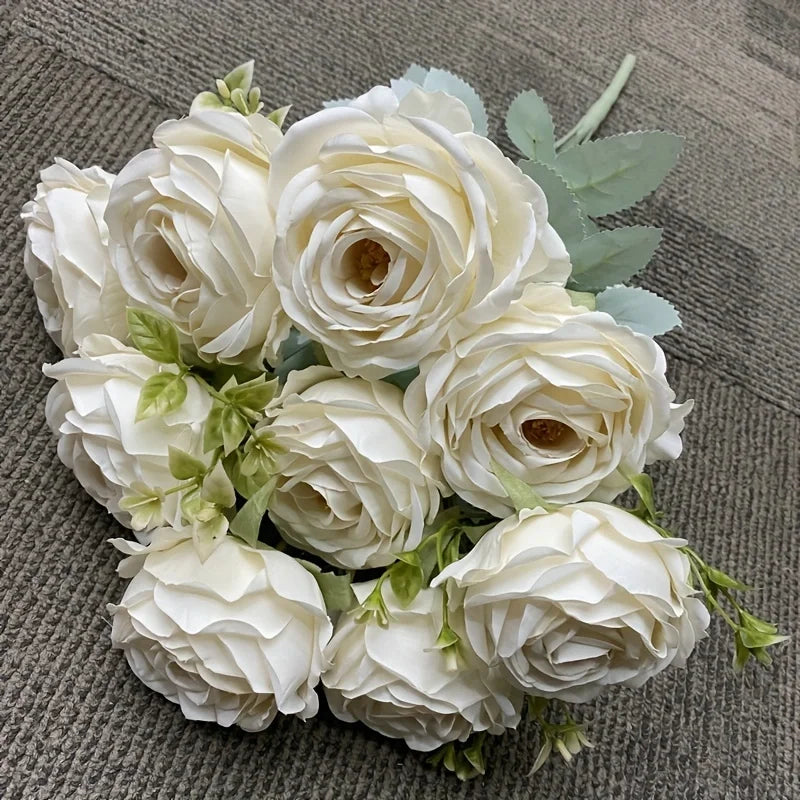 A bouquet of nine realistic white silk roses with green leaves arranged on a gray fabric.