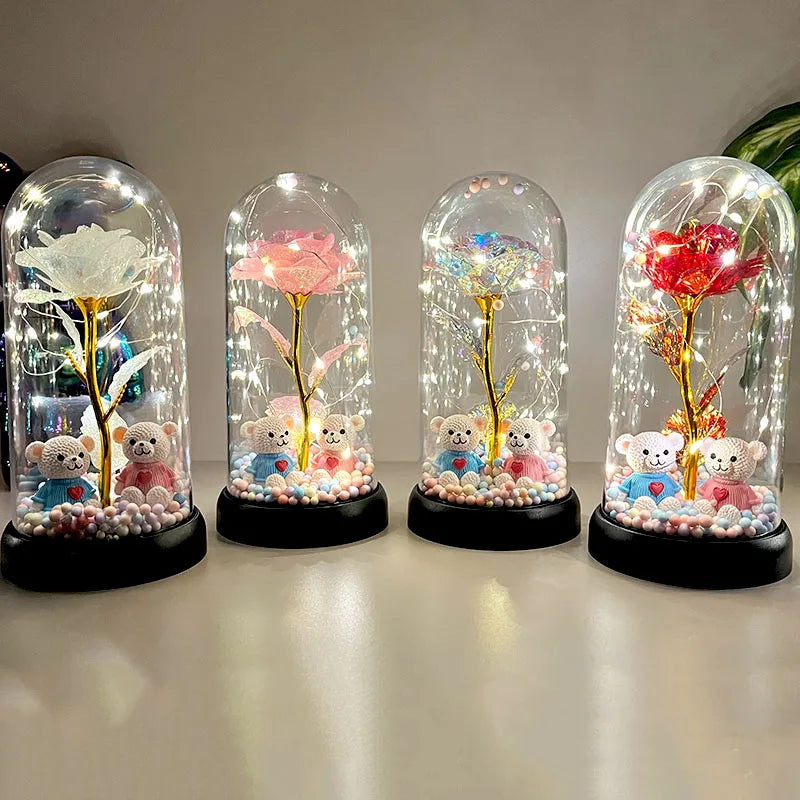 Cuatro flores de rosa de plástico en vidrio con ositos de peluche y elementos brillantes, exhibidas sobre una superficie blan