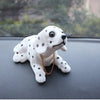 A white ceramic dog ornament with black spots and a gold chain is displayed on a dark surface.
