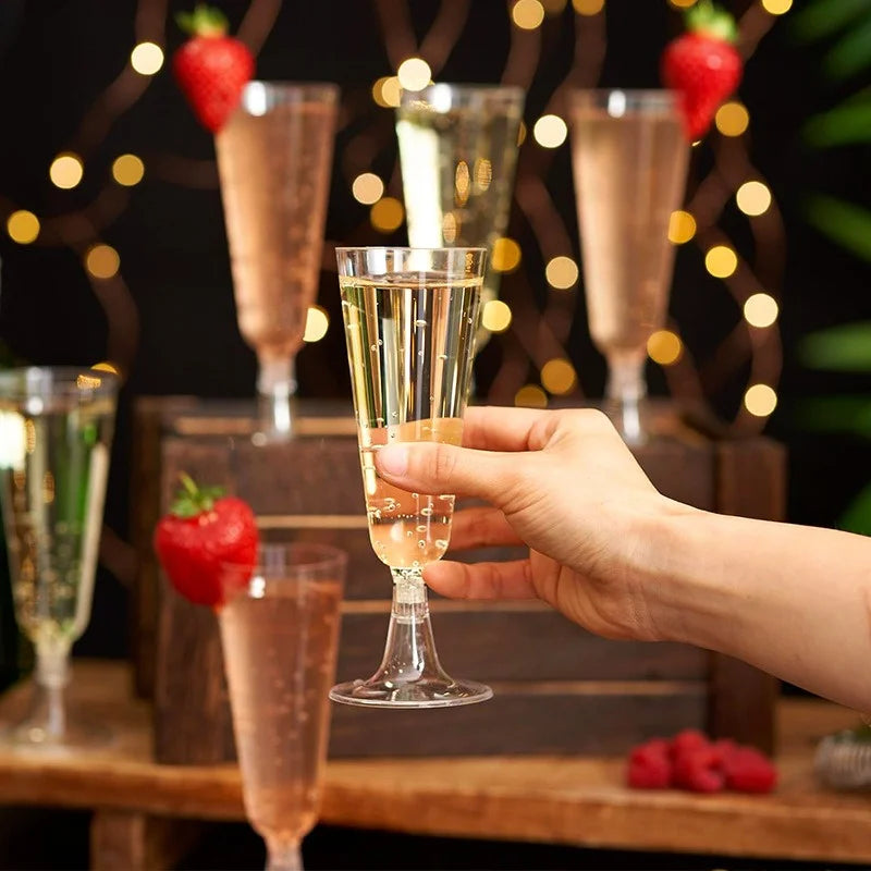 A hand holds an elegant disposable champagne glass filled with a light golden beverage against a dark background with bokeh l