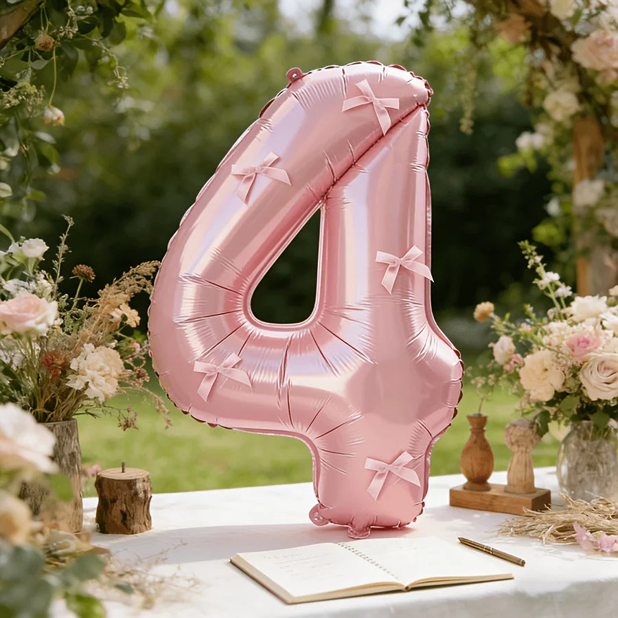 A pink foil number balloon shaped like the number 4 with a bow, placed on a white table surrounded by floral greenery.