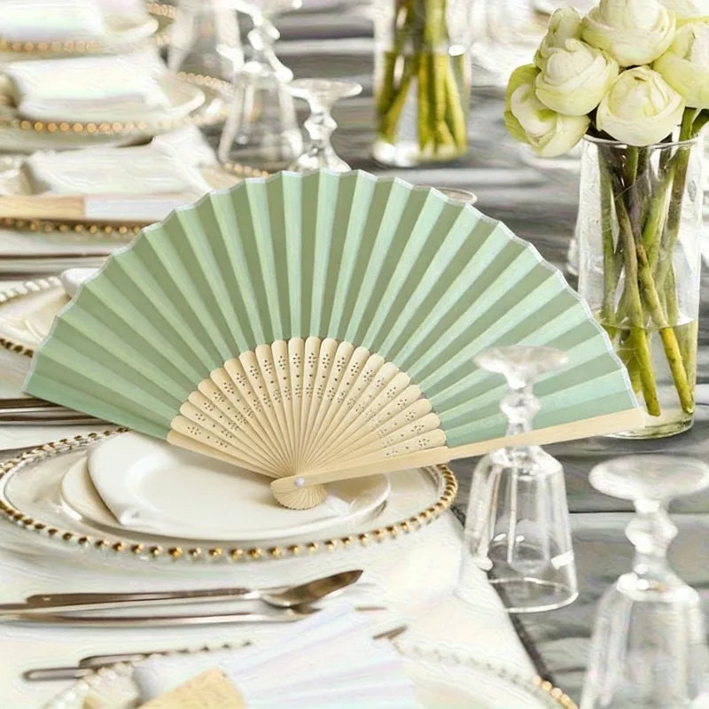 A set of light green folding hand fans displayed on a white table setting with glassware and floral arrangements.