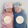 Two babies wearing knitted hats, one pink and one blue, with wooden plaques indicating it's a girl and a boy.