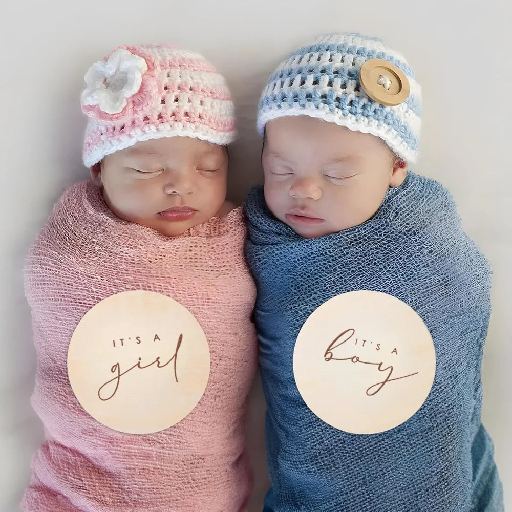 Two babies wearing knitted hats, one pink and one blue, with wooden plaques indicating it's a girl and a boy.