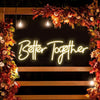A glowing neon sign spelling Better Together is displayed within a wreath of autumn leaves and flowers.