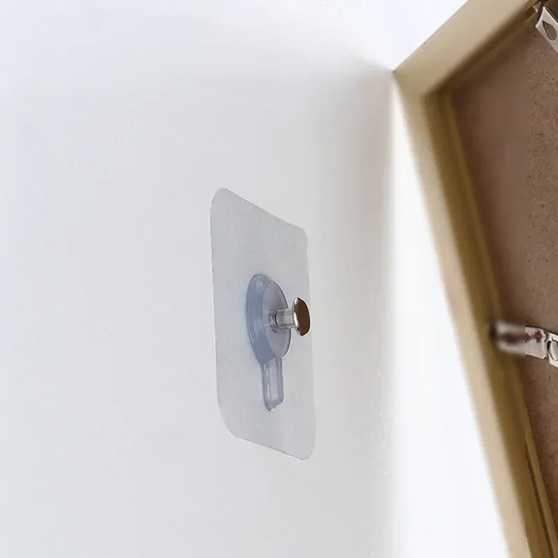 A transparent self-adhesive wall hook with a small metal loop is mounted on a white wall next to a picture frame.
