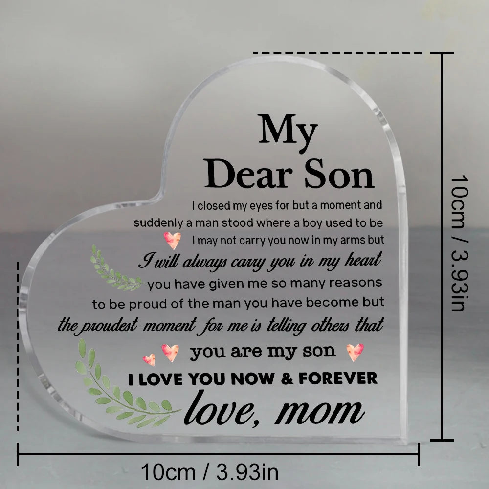 A clear heart-shaped acrylic keepsake plaque engraved with a message for a son, 'I love you now & forever, love, mom'.