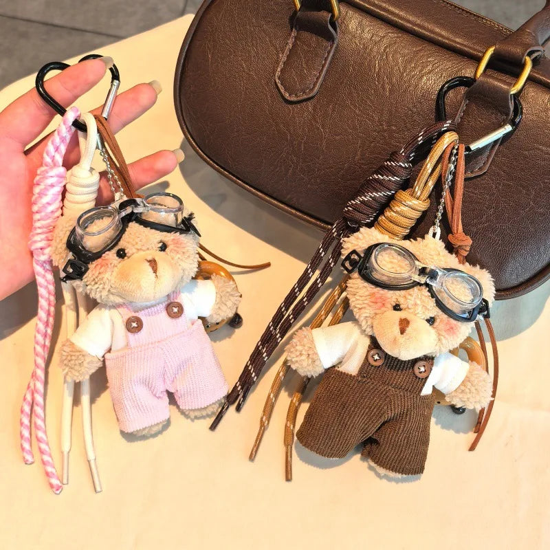 Un ours en peluche flocon portant des lunettes aviateur et des vêtements, à côté d'un sac en cuir marron.