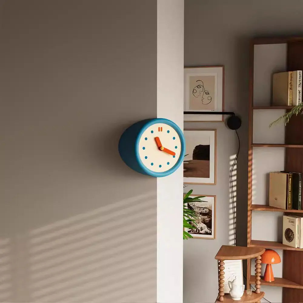 A modern blue resin wall clock is mounted on a beige wall next to a wooden shelf display.