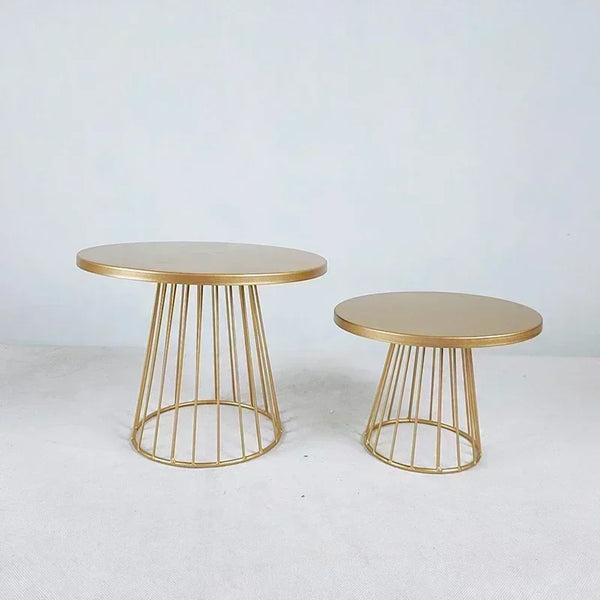 Two gold metal cake display stands with round tops and vertical wire bases on a white surface.