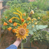 Una mano sostiene un ramo de flores de crochet hechas a mano en tonos amarillo y verde contra un fondo exterior desenfocado.