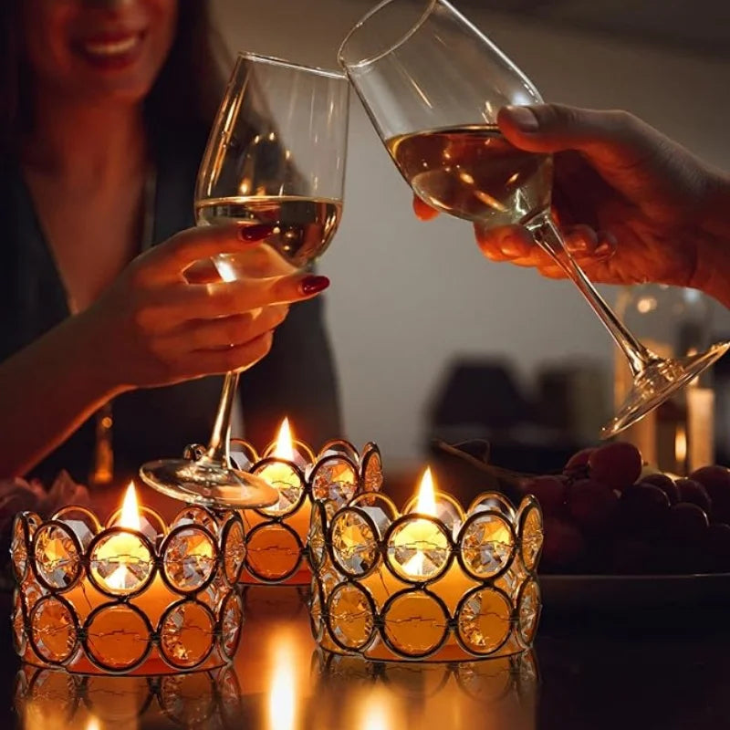 A hand holds a glass of amber liquid near glowing crystal tealight candle holders on a dark surface.