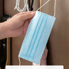 A hand holds a light blue pleated face mask hanging on a wall hook.