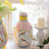 A pink and white striped ceramic vase with floral designs sits on a table next to a yellow candle.
