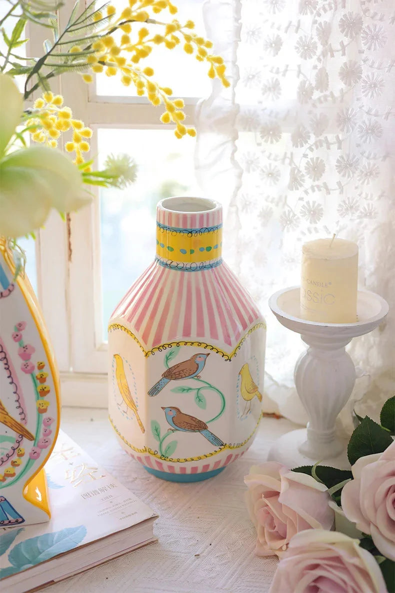 A pink and white striped ceramic vase with floral designs sits on a table next to a yellow candle.