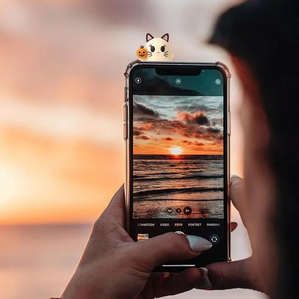 A hand holds a smartphone displaying a sunset over the ocean with a small cat charm decoration on the screen.