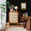 A wooden cabinet with drawers and a dark floral wallpaper background featuring a plant and a lamp.