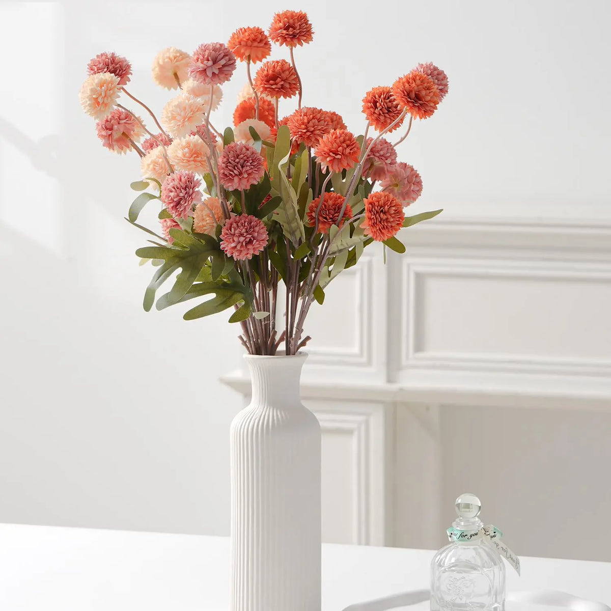 A bouquet of artificial chrysanthemum pompon flowers in shades of orange, pink, and peach displayed in a white vase.