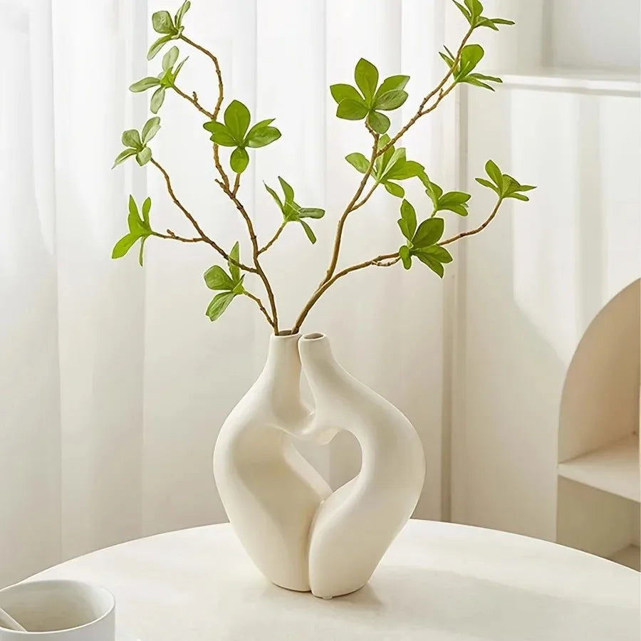 A white, modern heart-shaped vase holds a branch of green foliage in a bright indoor setting.