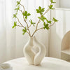 A white, modern heart-shaped vase holds a branch of green foliage against a bright background.